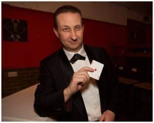 Professional close-up magician in a black tuxedo and bow tie holding the Ace of Diamonds towards the camera while smiling at an indoor event.